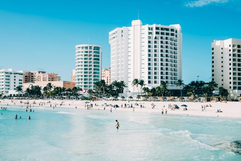 Esta imagen muestra una hermosa playa con aguas turquesas y arena blanca. En el fondo, hay varios edificios altos, probablemente hoteles, y palmeras que dan un toque tropical. Hay muchas personas disfrutando de la playa, ya sea nadando, caminando o tomando el sol. En el contexto de "LinkedIn México Consejos para Mejorar tu Marketing B2B", si tu empresa se dedica al turismo, el sector hotelero o servicios relacionados con la recreación en México, puedes utilizar imágenes de destinos tan atractivos como éste en tus publicaciones de LinkedIn. Compartiendo contenido sobre las ofertas turísticas, los servicios exclusivos de los hoteles o las oportunidades de inversión en el sector, y utilizando hashtags como #TurismoMéxico, #MarketingB2B y #HotelesMexicanos, podrás captar la atención de posibles clientes empresariales y mejorar tu estrategia de marketing B2B.